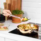En person laddar en FINMAT-sked med wokade nudlar och grönsaker, och visar upp dess djupa skål och robusta handtag designat för omrörning och servering.