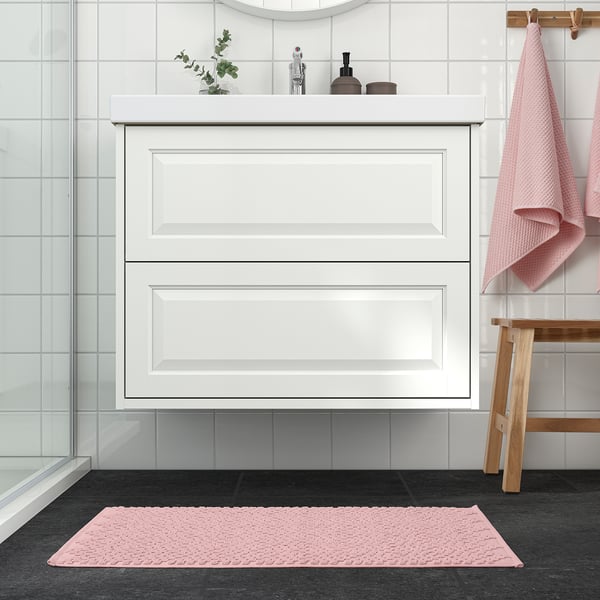 Modernt badrum med en ljusrosa FJÄLLKATTFOT badrumsmatta. Mattan passar snyggt framför det vita skåpet och kompletterar hängande handdukar och en liten växt.