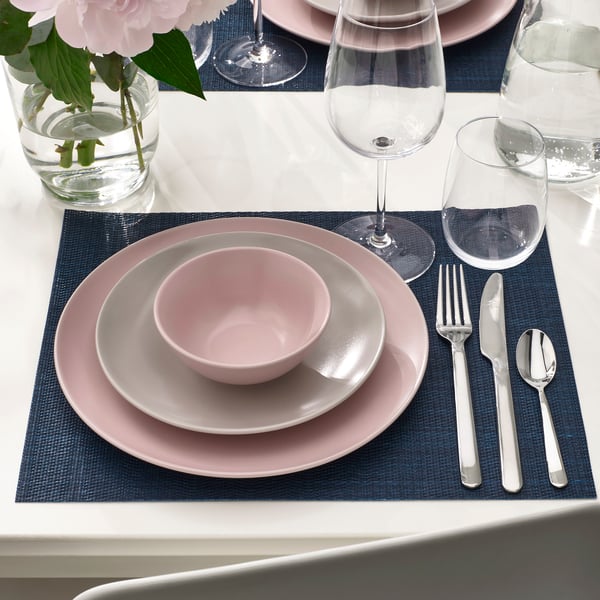 Dukning med rosa tallrikar, skålar och bestick på blå bordstabletter. Innehåller vinglas och en vas med rosa blommor.