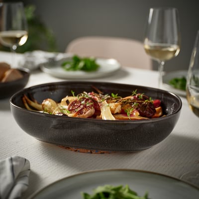 Mörkgrå ugnsform GLADELIG fylld med mat på ett matbord, tillsammans med vinglas och tallrikar med grönt, som serverar flera portioner.