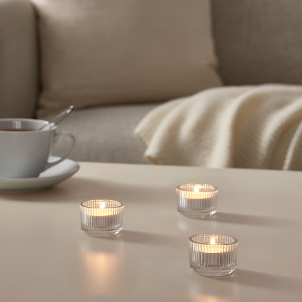 Tre tända GLIMMA-ljus på ett vitt bord, blå kopp, sked, beige filt på soffan, varm glöd.