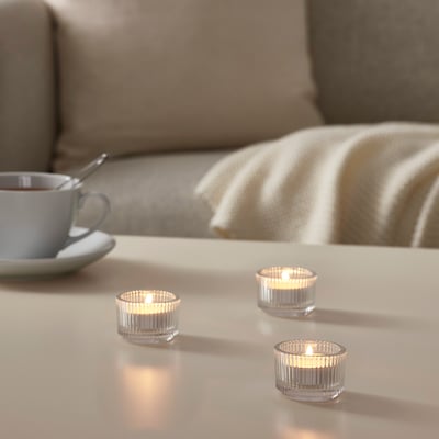 Tre tända GLIMMA-ljus på ett vitt bord, blå kopp, sked, beige filt på soffan, varm glöd.