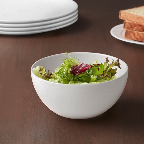 Vit skål med grön sallad på träbord, omgiven av tallrikar och bröd.