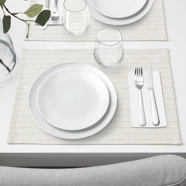 Elegant dukning med vit tallrik, glas och bestick på beige matta, redo för middag.