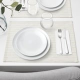 Elegant dukning med vit tallrik, glas och bestick på beige matta, redo för middag.