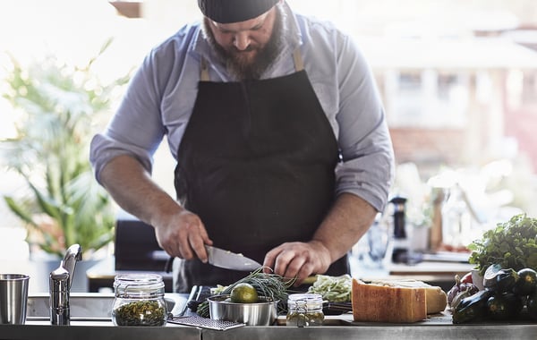 En skäggig kock lagar mat vid en disk med färsk frukt och grönsaker. Han är klädd i en grå skjorta och ett svart förkläde och skär upp ingredienserna med en kniv.