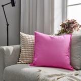 Ljusrosa GURLI-kudde på en grå soffa, texturerad med pom-poms, liten krukväxt i närheten, naturligt ljus genom fönstret, gardin i bakgrunden.