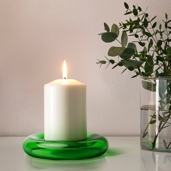 Ett tänt vitt ljus på en grön hållare i återvunnet glas. Bredvid den står en vas med löv som skapar en lugn atmosfär.