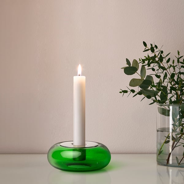 Ljusstake i grönt glas med tänt vitt ljus, bredvid vas med grönska.