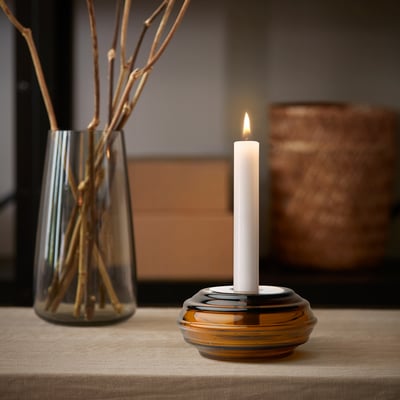 Brun glas HÄNGBJÖRK ljusstake med tänd ljus på träbord, bredvid en vas med grenar.