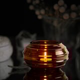En värmeljushållare i brunt glas, upplyst med ett stearinljus inuti, reflekterar varmt ljus, sitter på en yta bland suddiga dekorativa element.