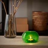 Litet grönt värmeljus i återvunnet glas i bubbelhållare, bredvid hög vas med kvistar.