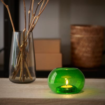 Litet grönt värmeljus i återvunnet glas i bubbelhållare, bredvid hög vas med kvistar.