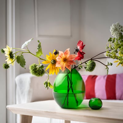 Grön vas av återvunnet glas HÄNGBJÖRK med blommor på träbord, två små gröna sfärer, vit/röd randig kuddebakgrund.
