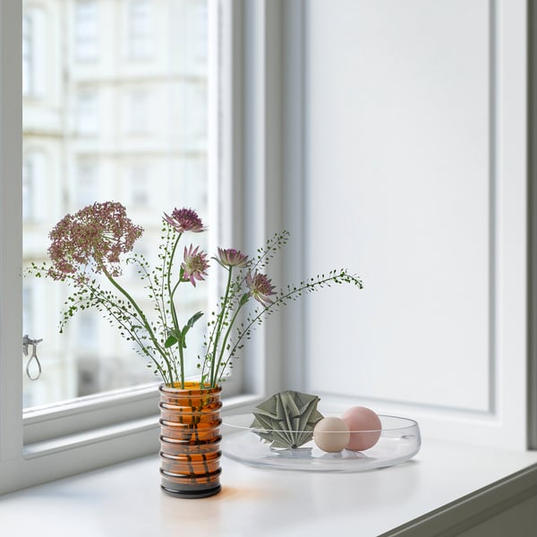 Vas på fönsterbrädan rymmer lila blommor, med hjälp av återvunnet glas. Bredvid den, en skål med sfärer och ett vikt föremål.