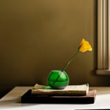 En liten grön glasvas rymmer en hög gul blomma som sitter på en trave med böcker.