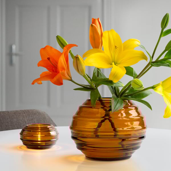 Brun glasvas som innehåller livfulla orange och gula blommor på en vit yta med ett tänt värmeljus i närheten.
