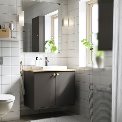 Modern grå badrumskommod med bänkskiva i ljust trä, vitt handfat, grå skåp, spegel och växter.