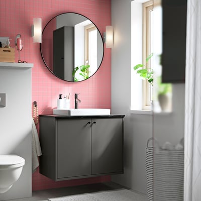 Modern grå HAVBÄCK kommod med rosa kakelväggar, rund spegel och vit diskho. Fönster och lampor ger naturligt ljus. Vit toalett & handdukshängare synlig.