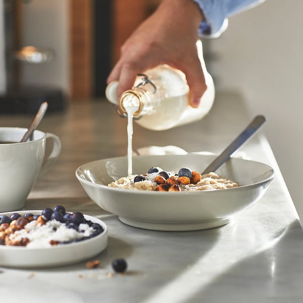 Personen häller mjölk i en skål med havregrynsgröt toppad med blåbär och nötter på bänkskivan, nära kaffekoppen.
