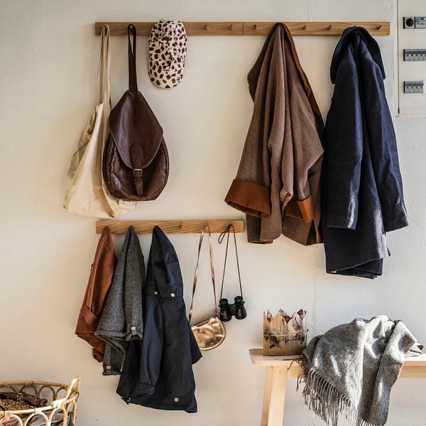En klädhängare i trä, HÖVOLM, med jackor, väskor och accessoarer. Den har en naturlig ekfinish och är monterad på en vägg.