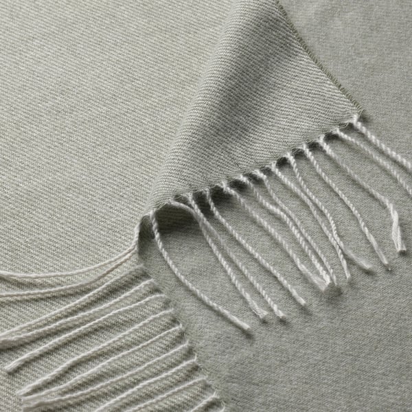 Mjuk grågrön vävd pläd med fransiga kanter, tillverkad av återvunnen polyester, draperad för att visa struktur.