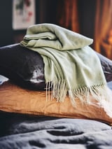 Ljusgrön HOLMVI pläd draperad på kuddar, mjuka, fransiga kanter.