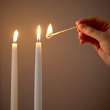 En hand tänder det sista av tre vita ljus, som hålls av tändstickor i ett svagt upplyst rum.