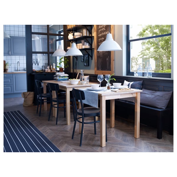 Modern matplats med ljust träbord, svarta stolar, pendellampor och kök i bakgrunden.