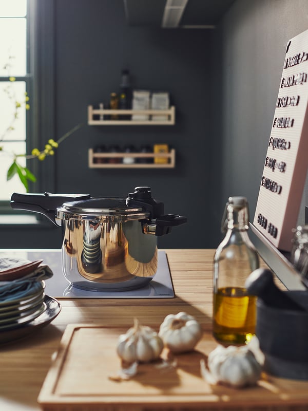 Modern köksbänk med IKEA 365+ tryckkokare i rostfritt stål, vitlök, oljeflaska och tallrikar, mörk vägg med hyllor.