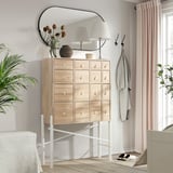 Modern ljus träbyrå med vita metallben, med flera små lådor, toppad med en lampa och dekorativa föremål.