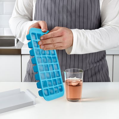 Person som håller i en blå iskubsbricka med 32 fack, som visar dess enkla utskjutningsfunktion. Glas dryck med is bredvid.