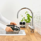 Person som tvättar grönsaker i diskbänken. Modern kromkran med sensor slår på och av vatten med handrörelse. Växter och kryddor i närheten.
