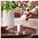 Person som tänder rosa JÄMNMOD doftljus på träbord bredvid vit vas med blommor.