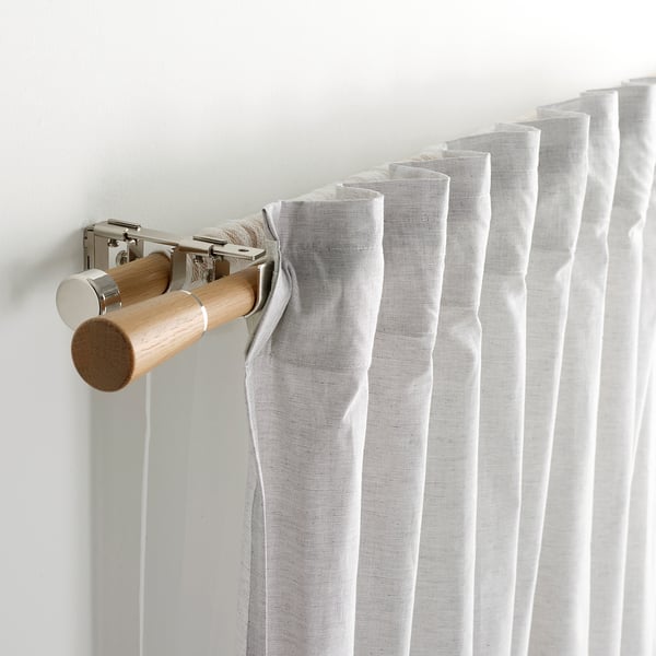 Närbild av gardinstång med ljust tyg. Visar stavar, ekknoppar och metallfästen.