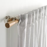 Närbild av gardinstång med ljust tyg. Visar stavar, ekknoppar och metallfästen.