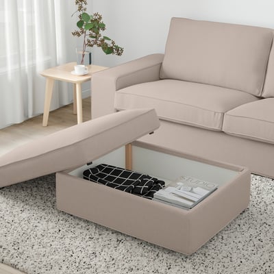 Beige KIVIK fotpall med förvaring, tidningar, filt, bredvid soffa och sidobord med vas och tekopp. Ljust, luftigt fönster.
