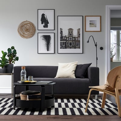 Modernt vardagsrum med grå soffa KLIPPAN, elegant soffbord, svartvitrutig matta, konstgallerivägg och trästolar.