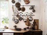 Kök med hängande kastruller och stekpannor; träbänk med växt och tillbehör; fönster med grönska.