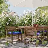 Uteplats med träbord, fyra stolar, blå kuddar, vitt paraply, glas och mat, omgiven av frodig grönska med vita blommor.
