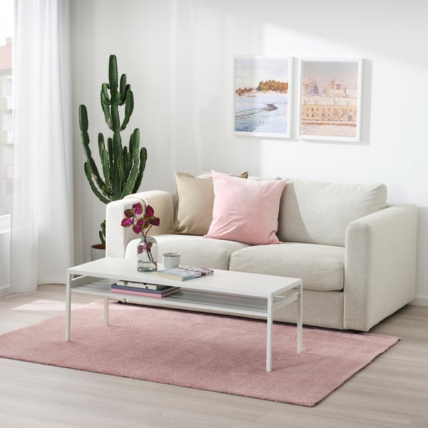 Minimalistiskt vardagsrum med KNARDRUP-matta, beige soffa, kuddar, soffbord med böcker och vas, kaktus, naturmålningar, fridfullt fönster.