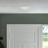KOPPLA vit taklampa, rund design, galvaniserat utseende, liten storlek men märkbar mot grön vägg, monterad ovanför vit dörr.