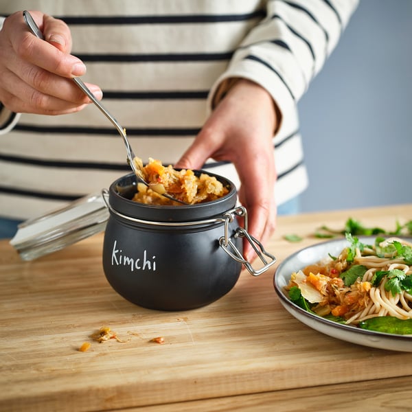 En person serverar kimchi från en svart KORKEN-burk märkt kimchi på ett träbord, bredvid en skål med nudlar och grönsaker.