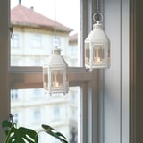 Två KRINGSYNT-lyktor i metall och glas hänger, varmt glödande bredvid växten.