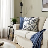 KRUTBRÄNNARE kudde, beige & blå, bomull, fyrkantig, tupp & blommig design. Mindre än stolen, bredvid en hög lampa.