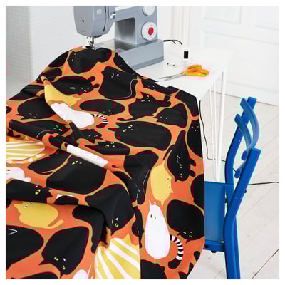 Symaskin med färgglatt katttryckstyg; orange, svart, gul; på vitt bord, blå stol bredvid.