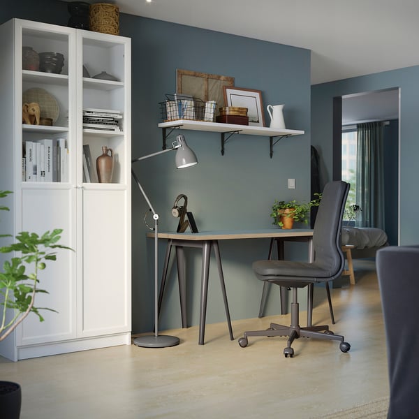 Hemmakontor med skrivbordet LAGKAPTEN: skiva i ljust trä, metallben, grå stol. Blå vägg, stark belysning, bokhylla vänster.