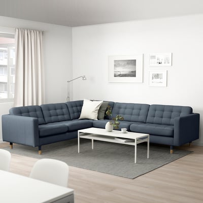 Modernt vardagsrum med l-formad sektionssoffa i blått tyg, vitt soffbord, golvlampa, matta och väggkonst.