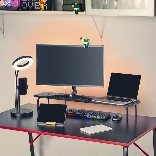 En spelmonitor står med tangentbord, mus och bärbar dator på ett svart skrivbord. LED-lampa och surfplatta i närheten.
