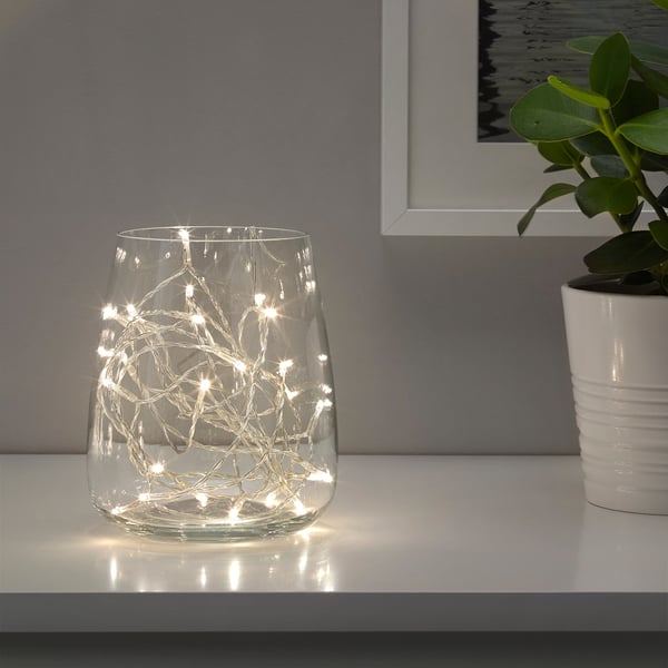 Vas i klart glas med varmvita LED-lampor som kastar ett festligt sken på en vit hylla.